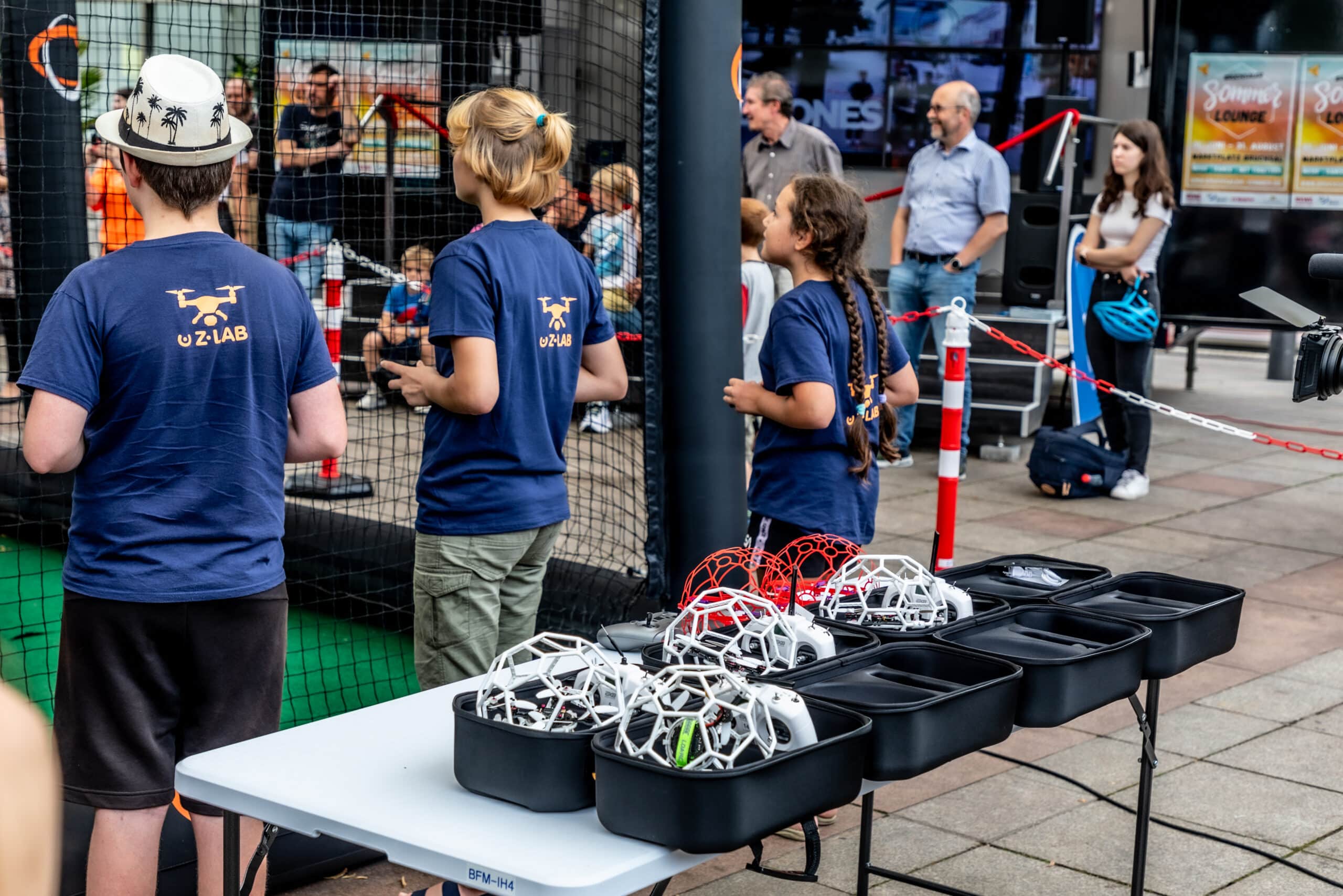Kinder stehen am Drohnenkäfig, im Vordergrund ist das Drohnenequipment für den Drone Soccer zu sehen. Im Hintergrund steht eine Menschenmenge, die das Geschehen verfolgt.
