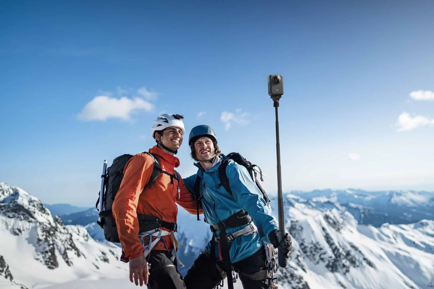 Two climbers filming themselves with a DJI Osmo 360 on an extension pole in front of snowy peaks